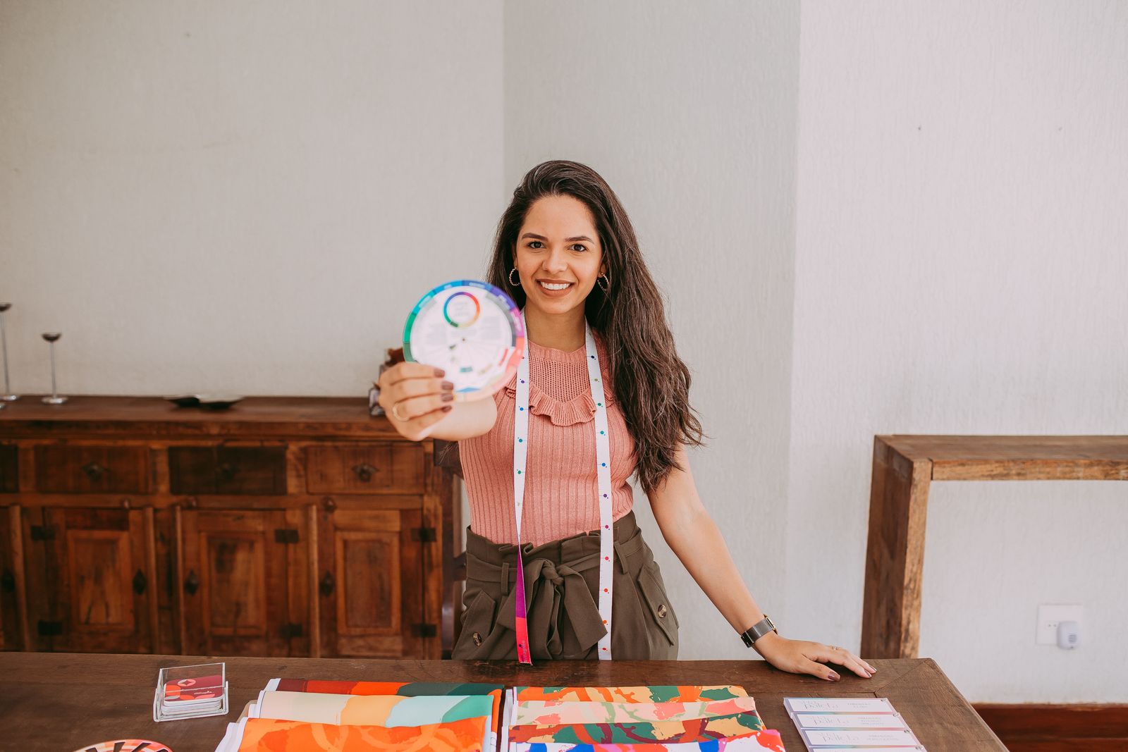 Artesã sorrindo atrás da bancada com tecidos estampados coloridos e círculo cromático na mão